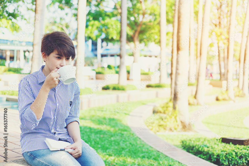 Beautiful short hair woman sitting on the floor and drinking a favorite beverage in white cup between her outdoor working business in the park. Beautiful short hair woman sitting on the floor and drinking a favorite beverage in white cup between her outdoor working business in the park.