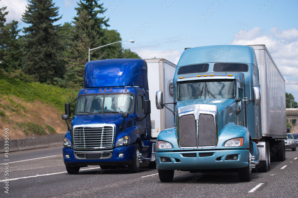 Two big rigs semi trucks in blue tone and different models with trailerd driving side by side Two big rigs semi trucks in blue tone and different models with trailerd driving side by side