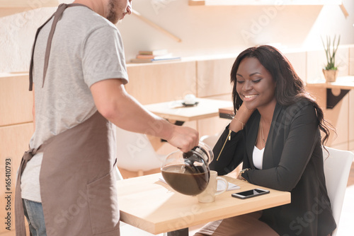 waiter pouring coffee for beautiful african american woman sitting in cafe