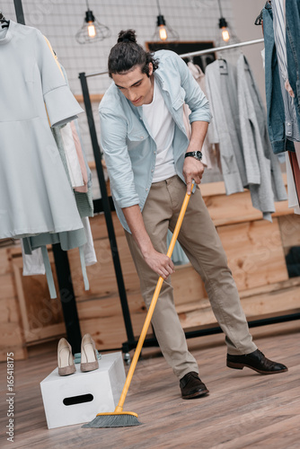 handsome young man with broom sweeping floor in shop before opening