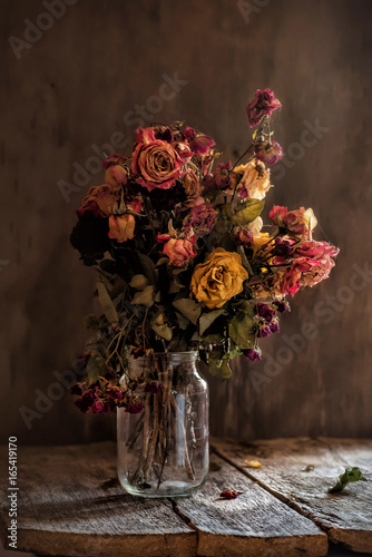 Bouquet of dried roses in a glass jar on a dark old wooden background. Vintage style hipster. Rough textures.
