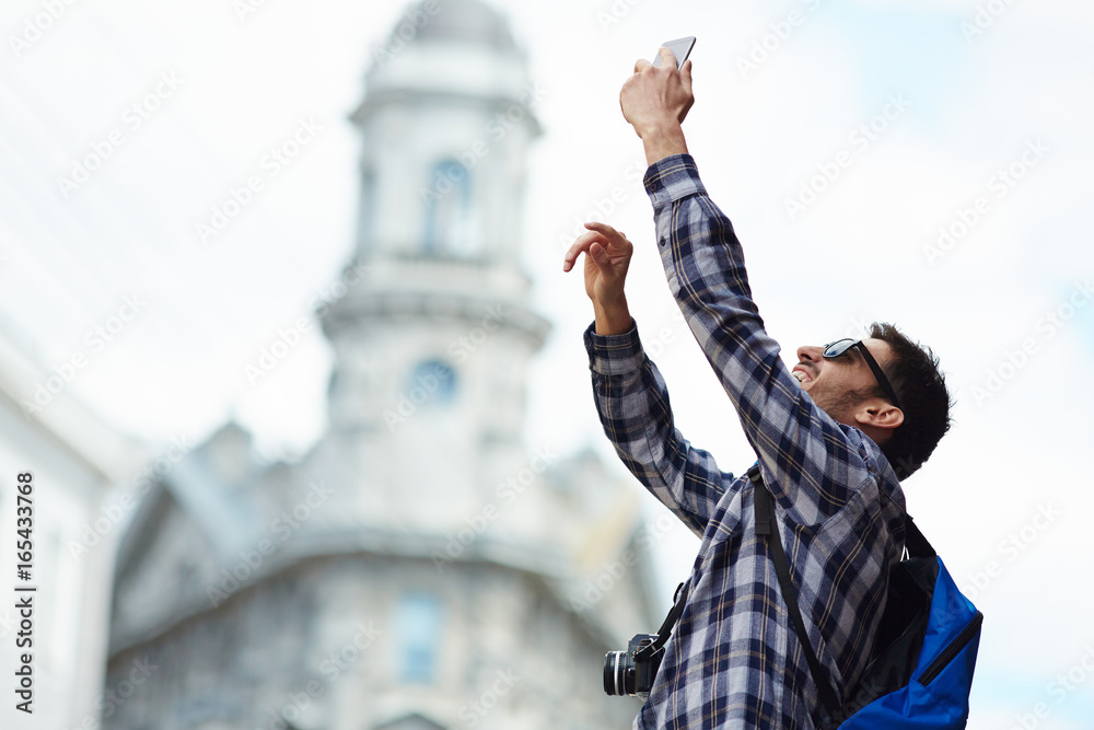 Fototapeta premium Portrait of modern young tourist on solo trip in Europe, happy man taking selfie photo in street of old city