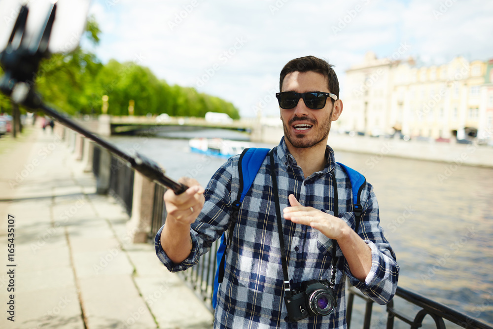 Portrait of handsome young man wearing sunglasses taking selfie photo using smartphone and monopod  on river bank enjoying tourist trip on vacation