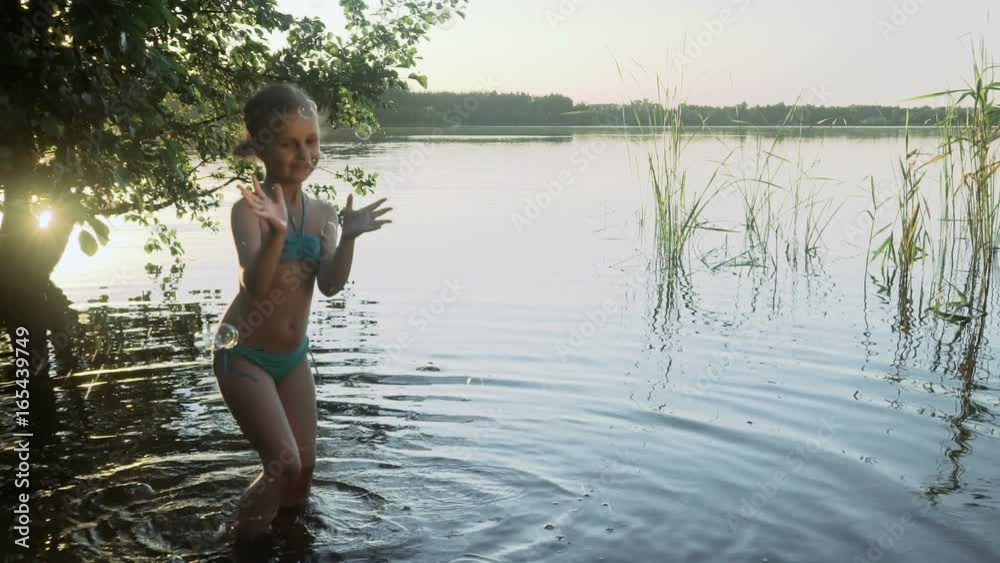Cute little girl is catching soap bubbles standing in the water of a picturesque forest lake. Slow motion.