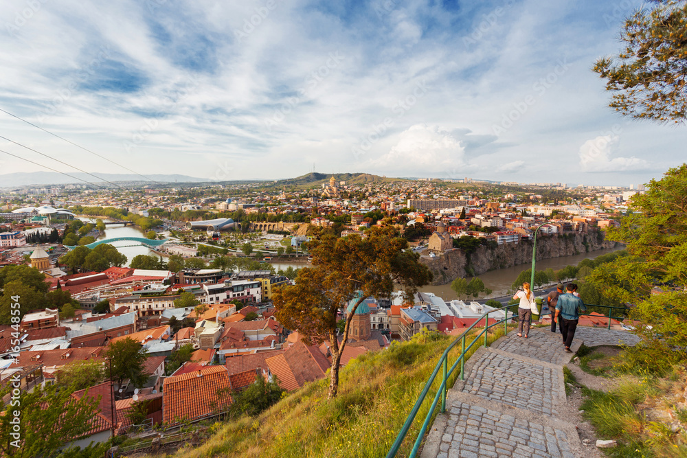 Panorama view of Tbilisi, capital of Georgia country. Tourists go down the stairs of Narikala fortress. Cable road above tiled roofs.