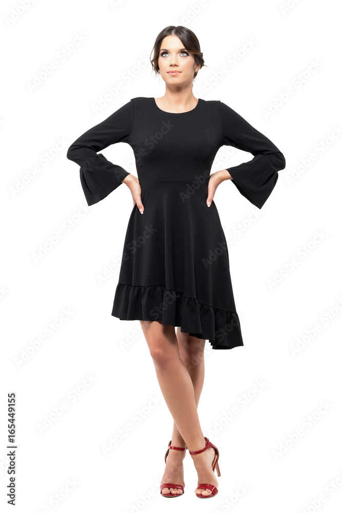 Gorgeous woman in black flounce dress and red high heels looking up with arms on hips. Full body length portrait isolated on white background. Gorgeous woman in black flounce dress and red high heels looking up with arms on hips. Full body length portrait isolated on white background.