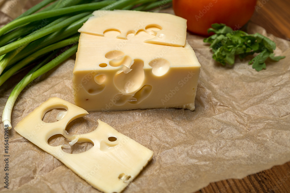 mouthwatering delicious piece of maasdam cheese and a slice on a wooden table surrounded by greenery and tomatoes