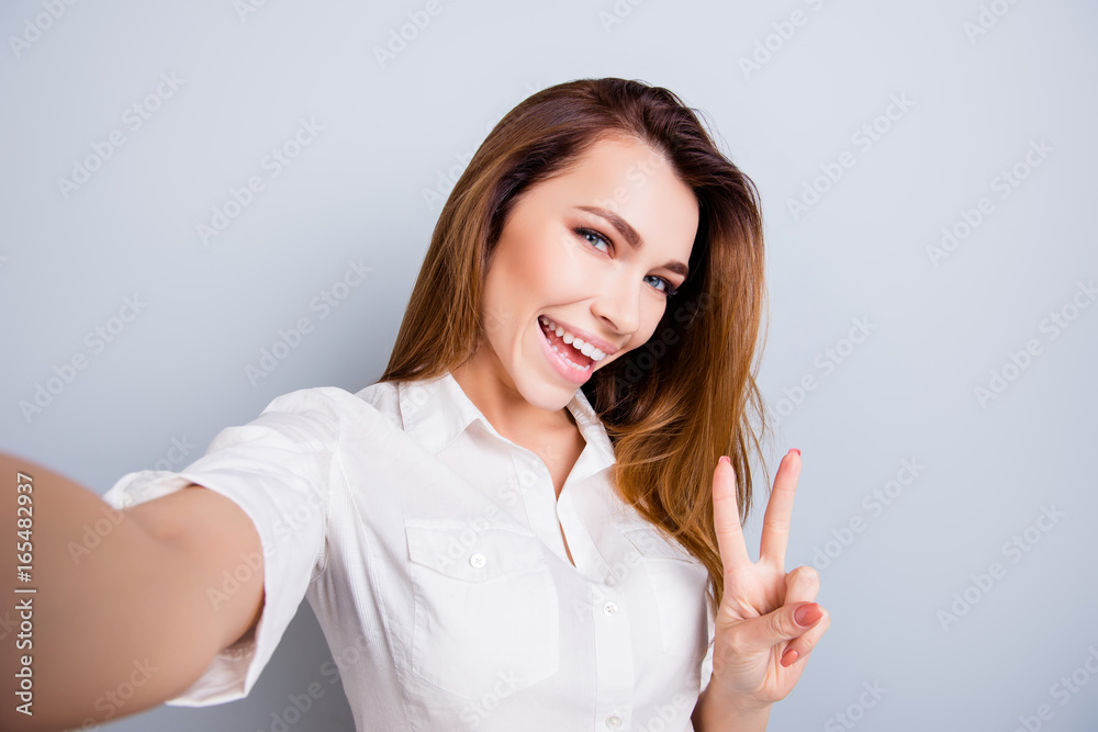 Selfie time! Funky mood. Attractive young lady is making a selfie on the camera, flirty and playful. Gesturing v sign, standing on pure light grey background Selfie time! Funky mood. Attractive young lady is making a selfie on the camera, flirty and playful. Gesturing v sign, standing on pure light grey background