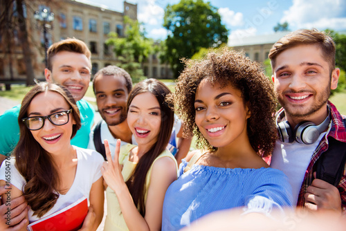 Selfie time! Six international students with beaming smiles are posing for selfie shot, african attractive lady is taking, outside school building. Gathered, cheerful, smart and successful youth