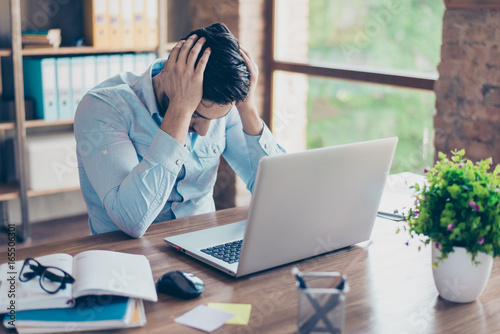 Close up portrait of tired sick young brunet man with strong migraine. He is wearing the formalwear, sitting at the workplace, holding his head, upset