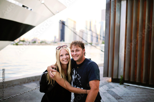 Young Traveling Couple Posing on Pier in Front of Ships and Sky Scrapers