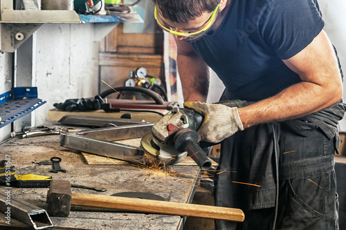 An adult man in work clothes and protective green glasses is engaged in manual labor and grinder metal an angle grinder on a wooden table in the workshop