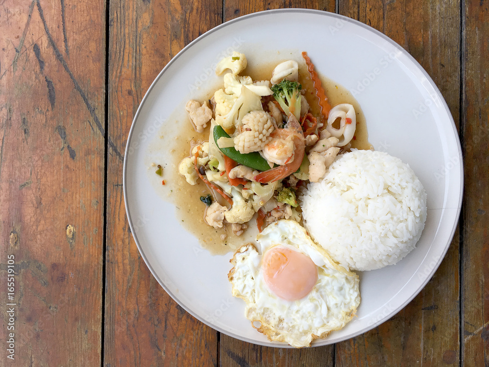 Mix of fried vegetables, Cauliflower, Carrot, Sugar Pea with seafood with rice and fried egg in white plate on wooden background. Thai style food. Mix of fried vegetables, Cauliflower, Carrot, Sugar Pea with seafood with rice and fried egg in white plate on wooden background. Thai style food.