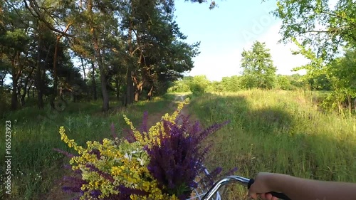 Woman riding a bike on a nature field forest with a bouquet of wild flowers in a basket in front of the wheel. Action camera go pro.