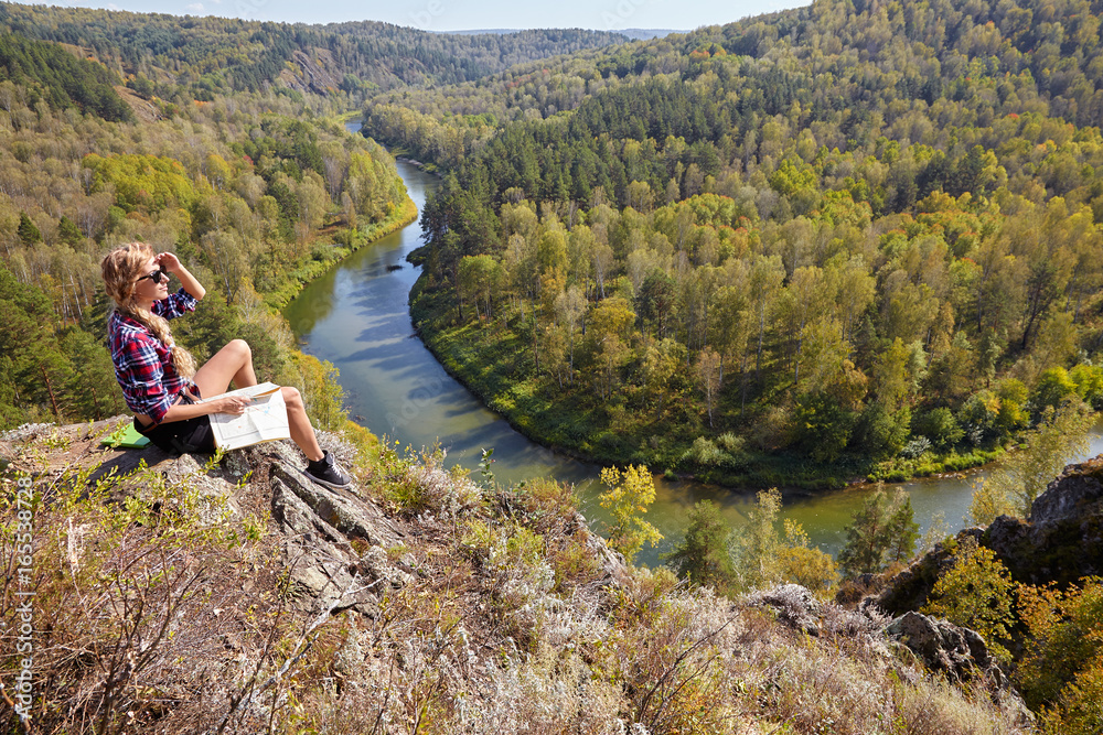 Young blonde woman tourist  sitting on a cliff with map in hands on background of siberian autumn landscape with the river Berd