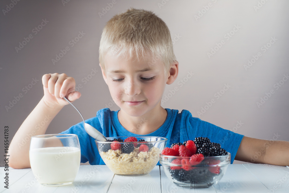 Little boy is eating oatmeal with berries. The concept of a healthy breakfast for children, porridge with raspberries and blackberries, a glass of yogurt on white wooden table. Little boy is eating oatmeal with berries. The concept of a healthy breakfast for children, porridge with raspberries and blackberries, a glass of yogurt on white wooden table.