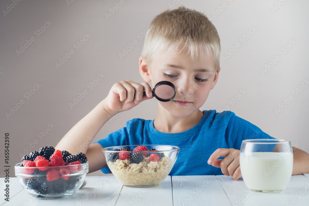 Boy examines a berries and oatmeal looking through a magnifying glass. The concept of a healthy breakfast for children, porridge with raspberries and blackberries, glass of yogurt on wooden table. Boy examines a berries and oatmeal looking through a magnifying glass. The concept of a healthy breakfast for children, porridge with raspberries and blackberries, glass of yogurt on wooden table.