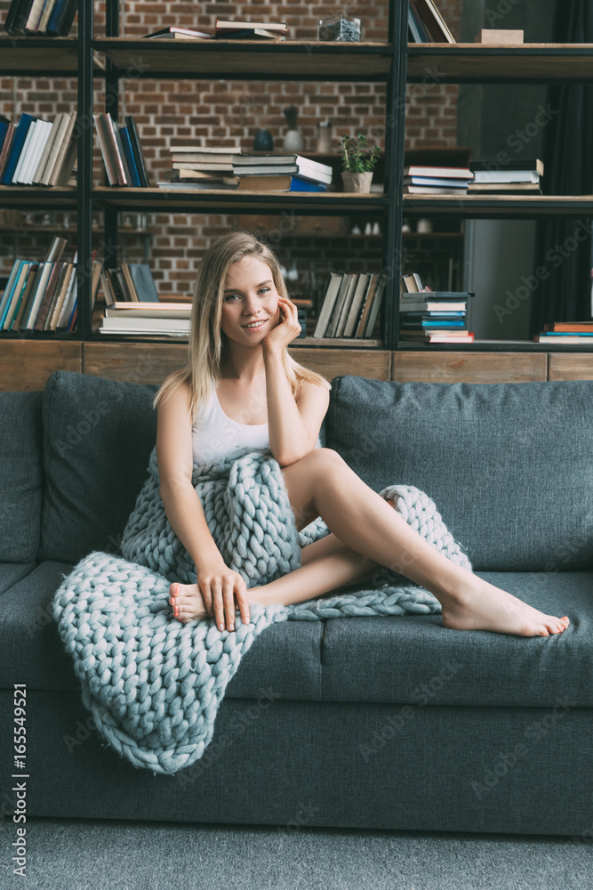 Obraz premium Young beautiful blonde woman wrapped in gray wool blanket on sofa in living room