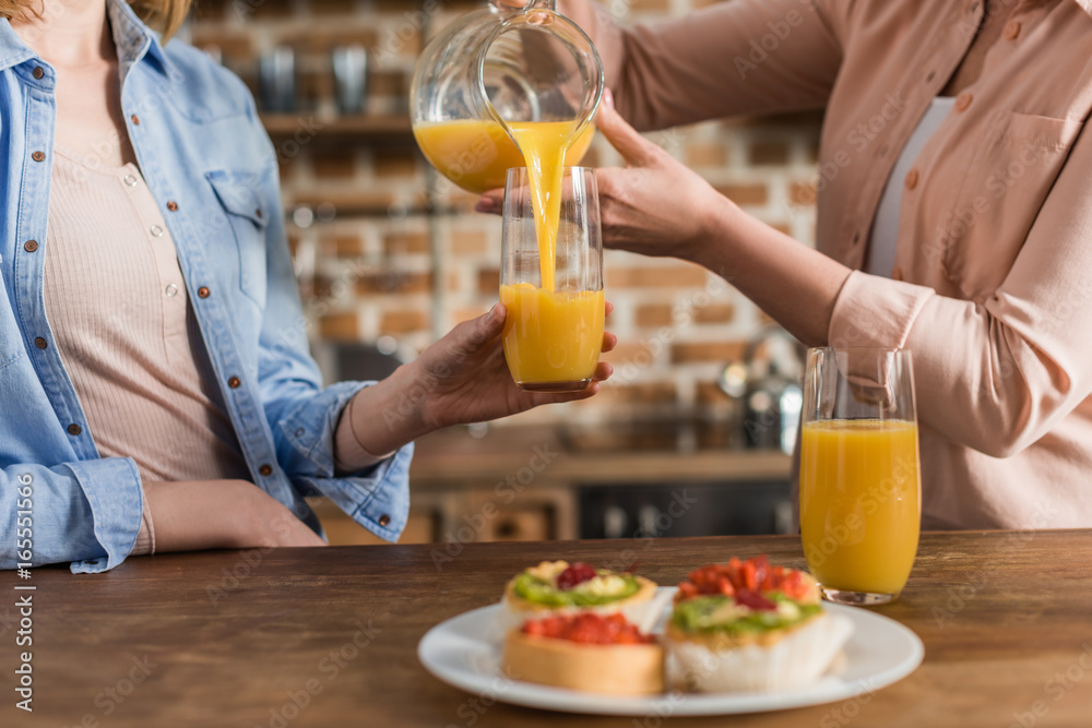 two women, senior and young drinking orange juice at table in kitchen. Woman pouring juice into glass two women, senior and young drinking orange juice at table in kitchen. Woman pouring juice into glass