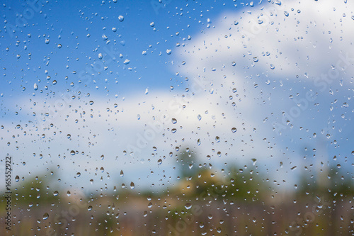 Raindrops on glass window next to blue sky and blurred green trees and white clouds. Summer weather concept.