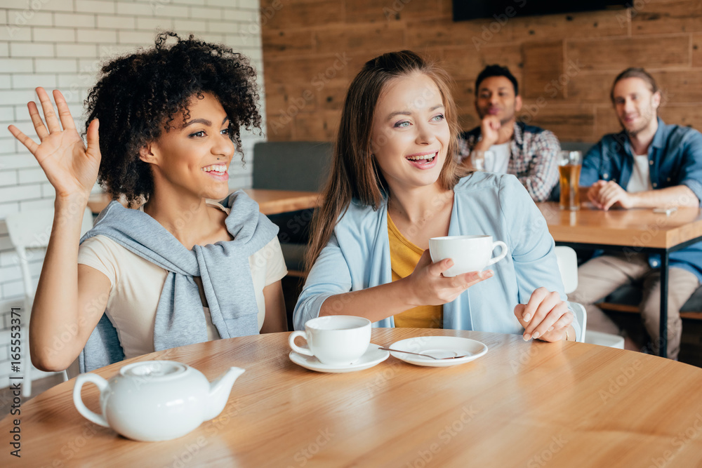 Two young women having tea in cafe and men sitting at next table looking at them