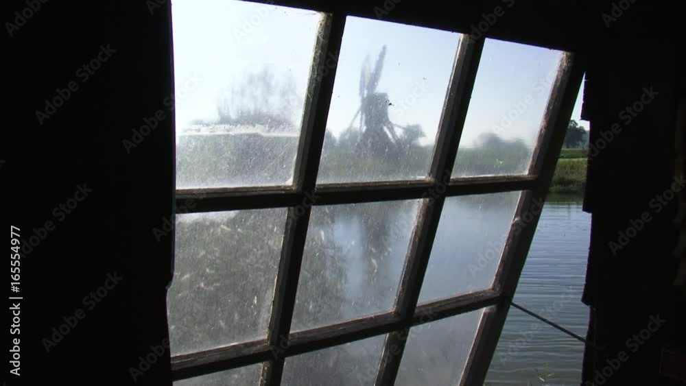 window view through a small divided window frame at a mill pond and small meadow mill outside. The meadow mill or spinnekopmolen serves as a drainage mill. window view through a small divided window frame at a mill pond and small meadow mill outside. The meadow mill or spinnekopmolen serves as a drainage mill.