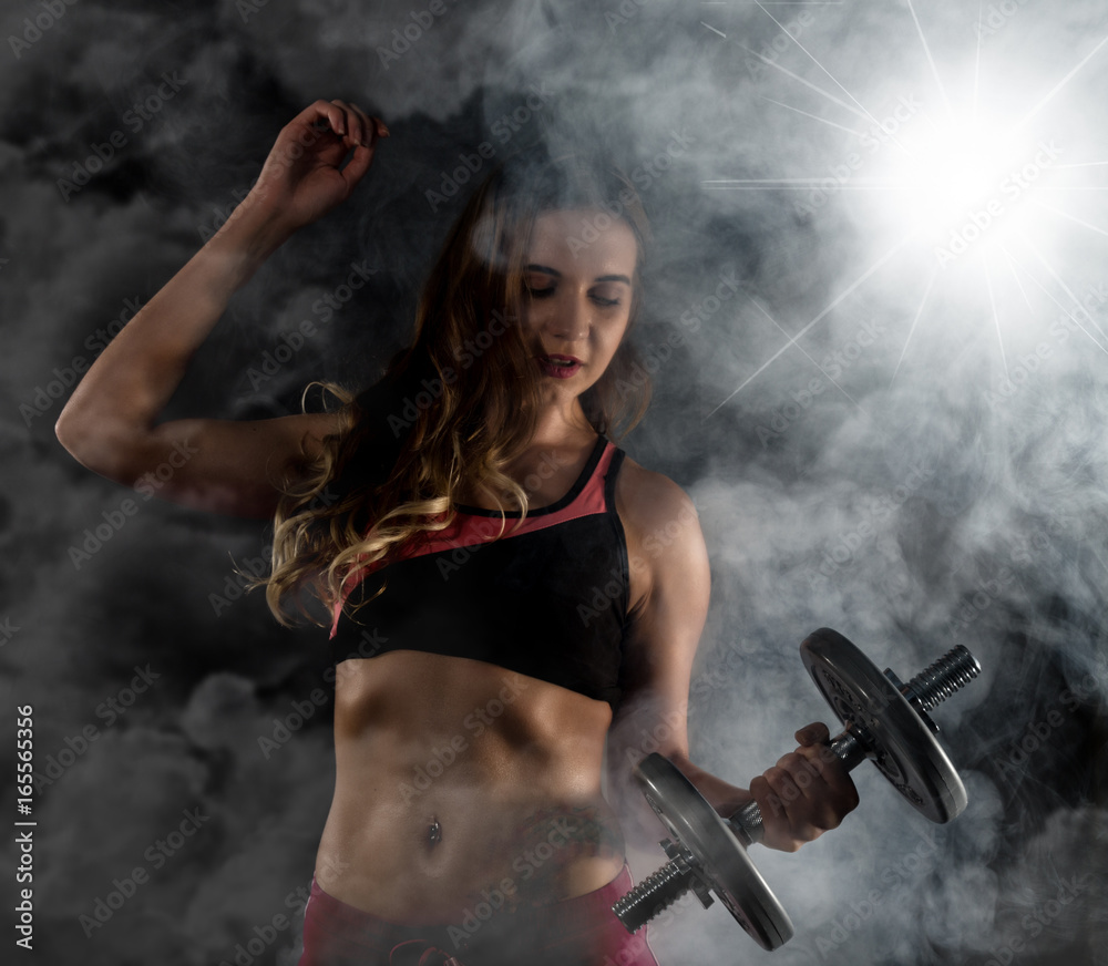 Fitness young woman in training with dumbbells, sporty muscular female brunette in smoke