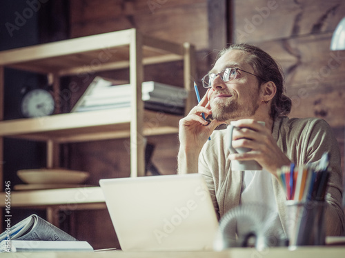Freelancer male at his working place smiling, thinking. Young man sitting at wooden table, holding mug and dreaming.