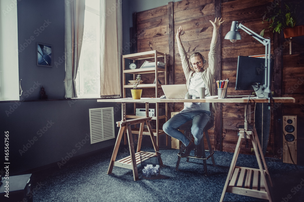 Freelancer man at his home office. Young man working online streching hands up.
