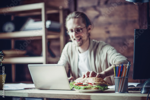 Man working at home going to eat at working place. Freelancer drawing hand to grab sandwich.