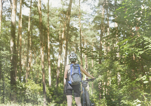 девушка в шлеме с рюкзаком и велосипедом стоит в летнем лесу солнечным днем