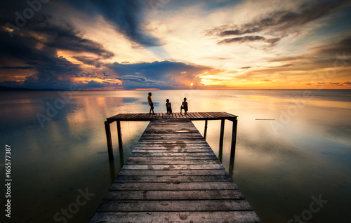 Wooden jetty during sunset with reflection. three unknown Sihleoutte of  kids appear on the image.  soft focus due to long expose.