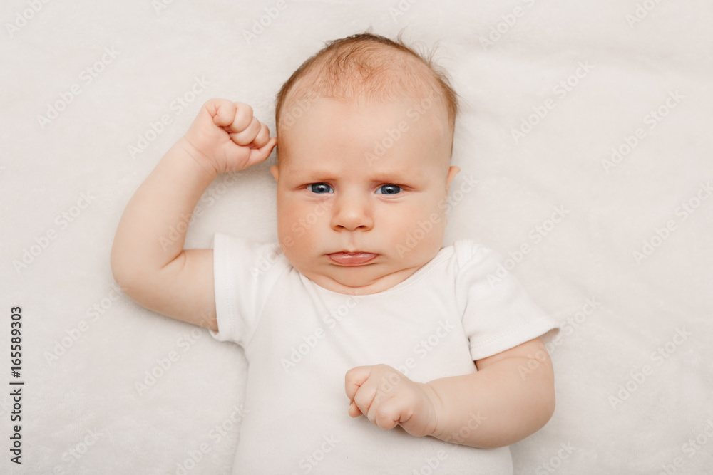 Closeup portrait of adorable funny white Caucasian baby with blue grey eyes lying on bed. Aware cute newborn on white background in studio. Little surprised child looking in camera making faces.