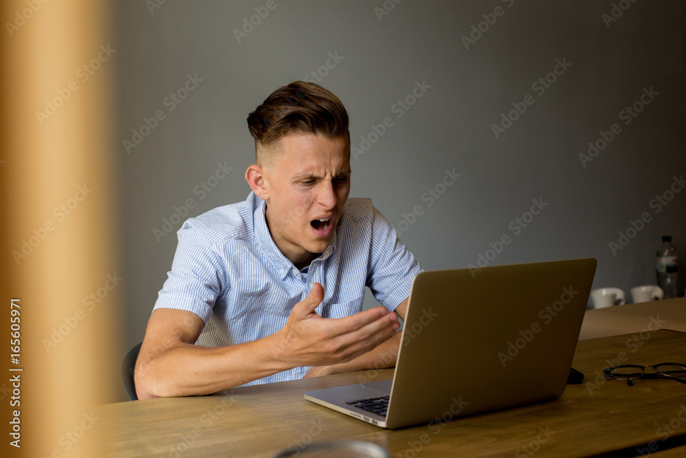 Confused and annoyed young man student shows his frustration by gestures towards computer and information on screen on social media Confused and annoyed young man student shows his frustration by gestures towards computer and information on screen on social media