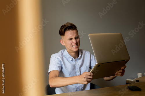Very angry and annoyed young man slams expensive technology laptop on table, because does not understand information on screen, typical internet meme