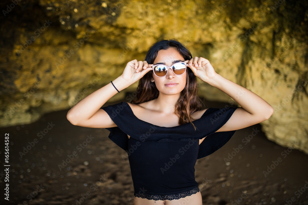Happy beautiful latin woman wearing sunglasses smile near on rocks beach on summer vocation time Happy beautiful latin woman wearing sunglasses smile near on rocks beach on summer vocation time