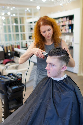 Girl hairdresser with curly red hair cuts young, handsome guy in a beauty salon