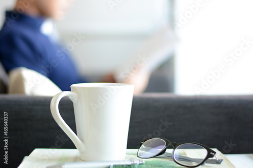 Coffee cup, eye glasses & pen over a book with blur background of a man reading book