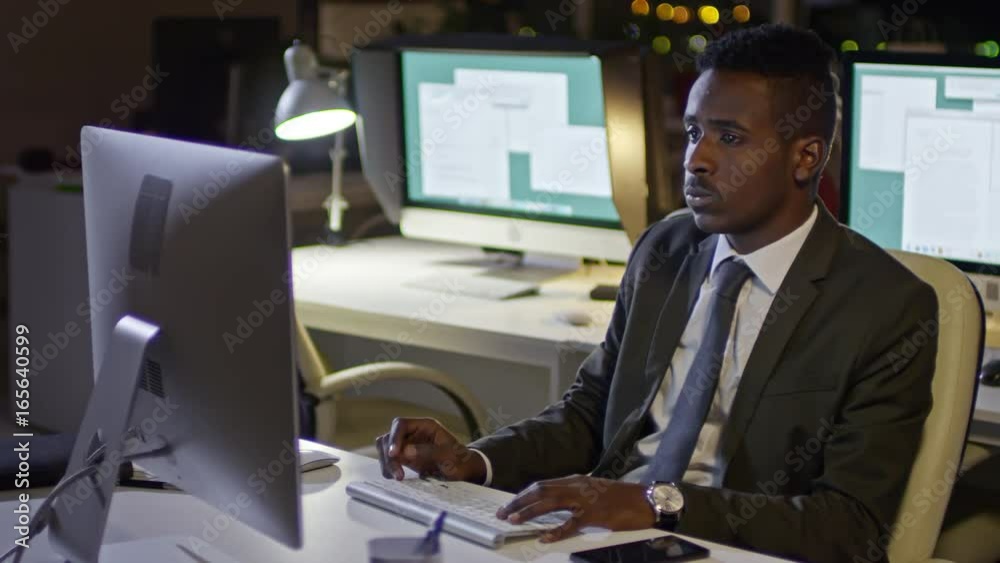 Tracking shot of young African businessman typing on computer and talking on the phone while working late in office Tracking shot of young African businessman typing on computer and talking on the phone while working late in office