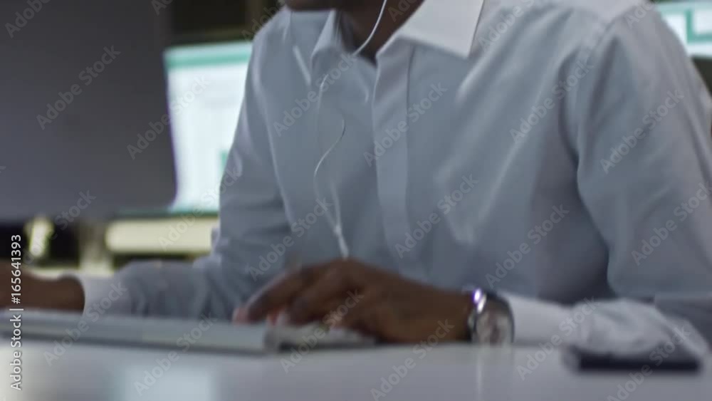African office worker listening to music with earphones, typing on computer and singing; handheld camera moving down to keyboard and up to face