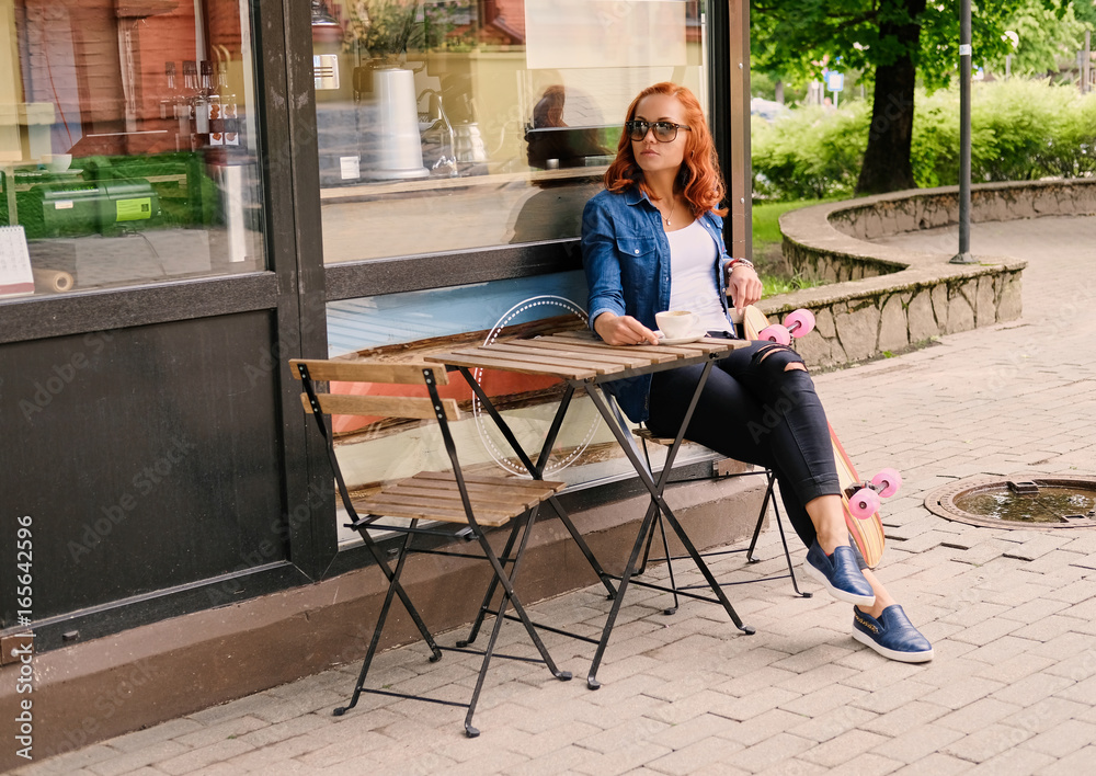 Redhead female drinks coffee at the table in a cafe on a street.