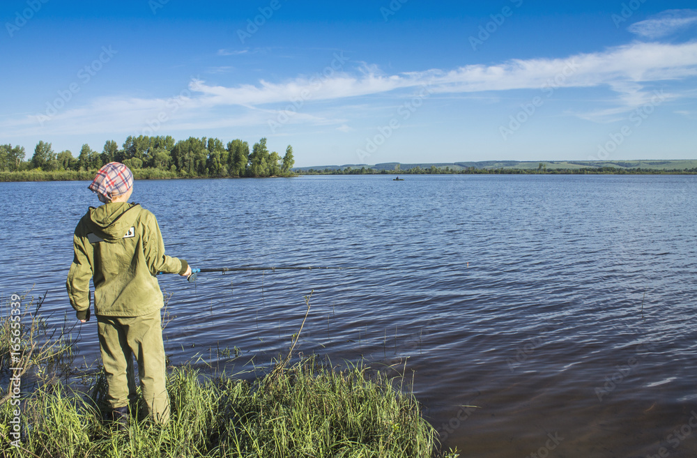 A young woman in green clothes is standing on the river bank and fishing with a fishing rod.