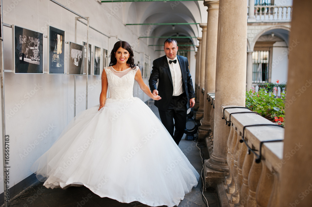 Naklejka premium Amazing young attractive newly married couple walking and posing in the downtown with beautiful and ancient architecture on the background on their wedding day.