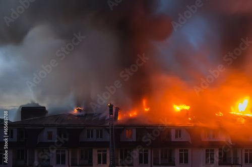 Burning fire flame with smoke on the apartment house roof in the city, firefighter or fireman on the ladder extinguishes fire