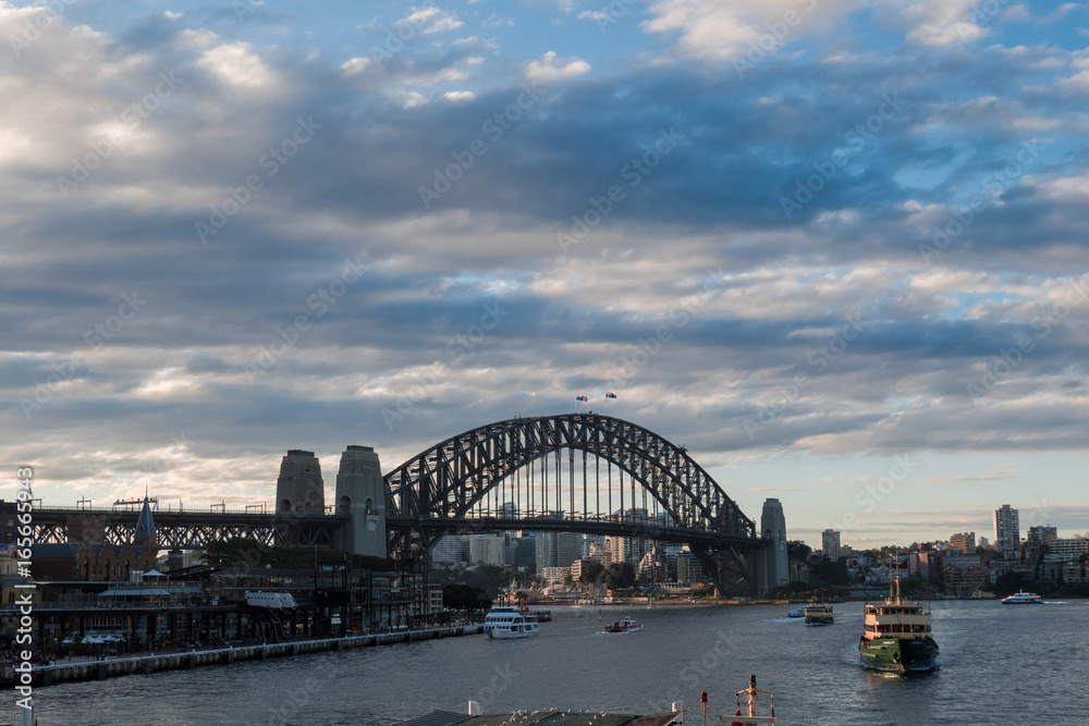 A cloudy view of Sydney Harbour Bridge with few ferries in the harbour area.