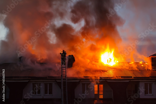 Burning fire flame with smoke on the apartment house roof in the city  firefighter or fireman on the ladder extinguishes fire