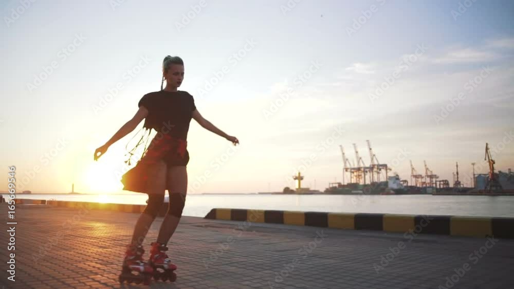 Young stylish funky girl with green hair riding roller skates and dancing near sea port during sunset, slow motion
