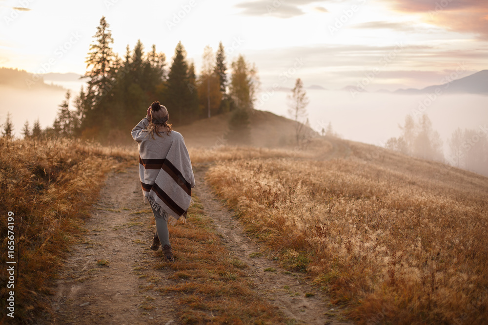 Young woman over the clouds in the valley looking at calm sunrise. Hiker girl wrapping in warm poncho outdoor. Successful woman hiker enjoy the view on mountain top.