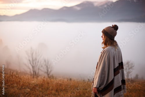Young woman over the clouds in the valley looking at calm sunrise. Young girl over the clouds. Wonderful landscape with cloud inversion. Successful woman hiker enjoy the view on mountain top. © Serhii