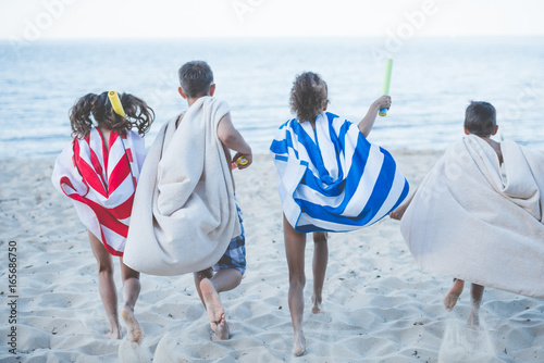 back view of children with towels as superhero robes and water toys in hands running on beach photo